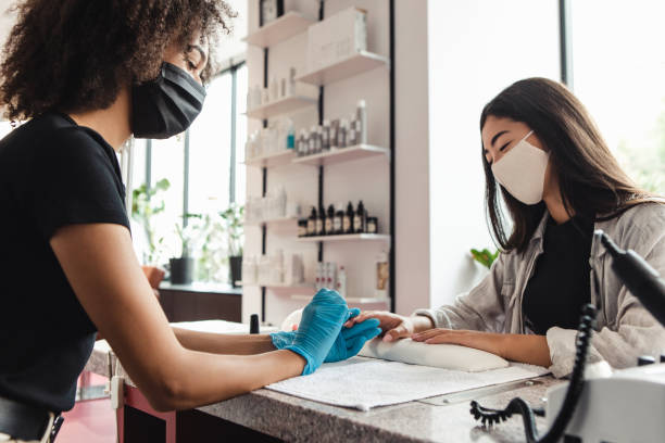 Manicure master in protective medical mask in beauty salon. African american girl in gloves doing nail procedure Asian woman and manicure master in protective medical mask in beauty salon. African american girl in gloves doing nail procedure in interior manicure stock pictures, royalty-free photos & images