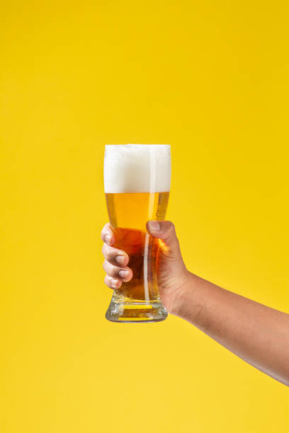 el brazo del hombre sostiene un vaso de vidrio con cerveza en su interior, espuma blanca y un fondo amarillo sólido - mano agarrando botella de cerveza y taza fotografías e imágenes de stock