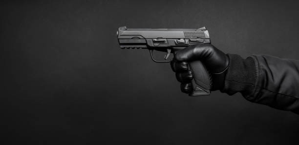 A man's hand in a black leather glove holds a pistol. Weapon in the hand of a man on a dark background. Weapon threat. stock photo