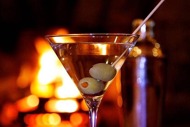Martini by the Fire Close-up of Martini with two olives with shaker and roaring fire in background  dirty martini stock pictures, royalty-free photos & images