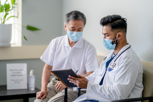A masked senior Asian man is seated next to a doctor for a medical check up appointment. The male doctor is holding a digital tablet and showing his patient the results of his medical tests.