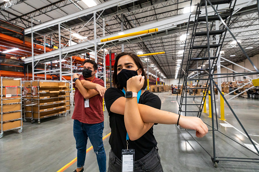 A young man and woman wearing face masks working in a massive fulfillment center, doing gentle stretching exercises to ensure the health and wellbeing of the employees.