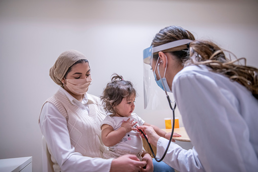 A masked female doctor is using a stethoscope to listen to a toddler girl's chest. The young patient is sitting on her Muslim mother's lap at the medical office.