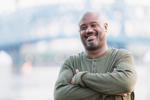 A mature African-American man in his 40s standing on a city waterfront smiling confidently at the camera, arms folded. The water and a bridge are out of focus in the background.