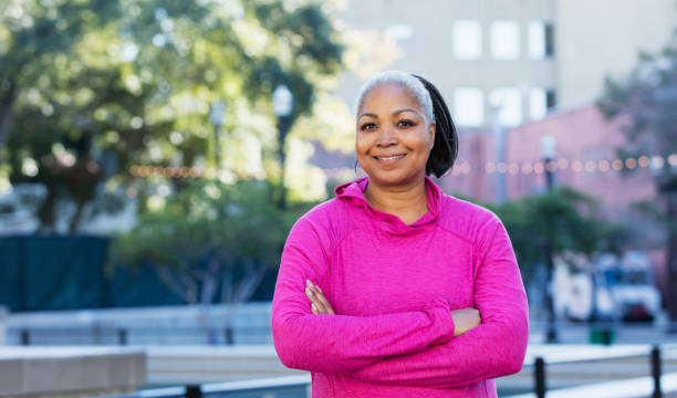 Mature African-American woman outdoors in city A mature African-American woman in her 50s wearing a pink hooded sweatshirt, smiling as she stands outdoors in the city, a tall building out of focus in the background. voluptuous women images stock pictures, royalty-free photos & images