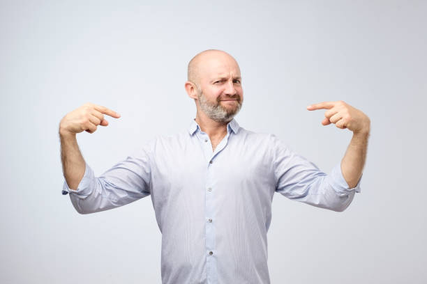 Mature bald adult man with beard standing over grey grunge wall looking confident with smile on face. Mature bald adult man with beard standing over grey grunge wall looking confident with smile on face. He is selfish a little and is pointing with fingers at himself one man only stock pictures, royalty-free photos & images