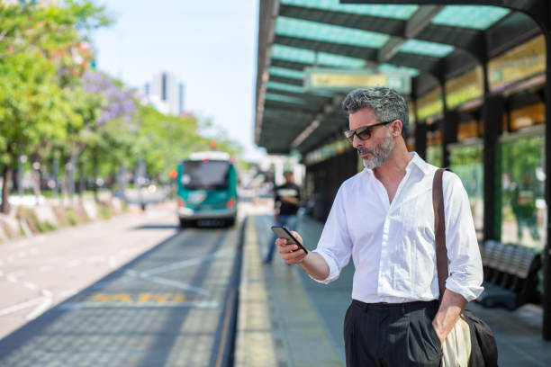 homme d’affaires mûr dans la gare routière utilisant le téléphone intelligent pour vérifier l’horaire de voyage - smartphone car photos et images de collection