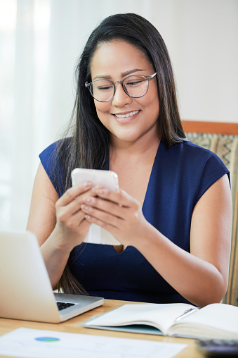 Portrait of smiling mature businesswoman working at office desk and answering text messages on smartphone
