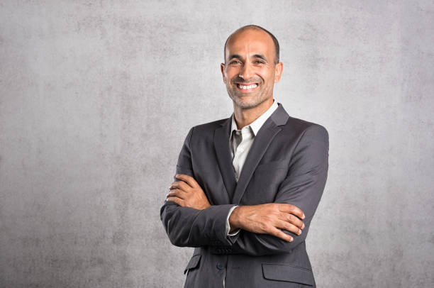 Mature confident businessman Portrait of happy mature businessman smiling on grey background. Successful senior leader in formal standing against grey wall with crossed arms. Satisfied hispanic man feeling proud and looking at camera. employee photos stock pictures, royalty-free photos & images