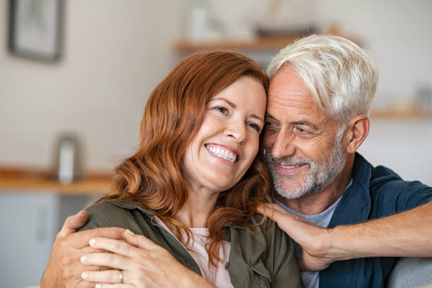 Mature couple cuddling in love Cheerful mature couple embracing while sitting on couch. Senior man giving woman a hug while looking at her with love. Loving old husband embracing from behind smiling wife at home. mid adult couple stock pictures, royalty-free photos & images