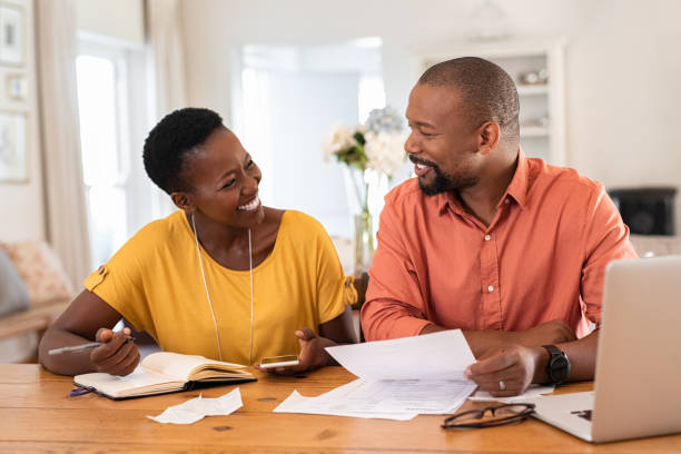 Mature couple managing home finance Cheerful mature couple sitting and managing expenses at home. Happy african man and woman paying bills together and managing budget. Black smiling couple checking accountancy and bills while looking at each other. bank account stock pictures, royalty-free photos & images