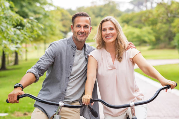 Mature couple riding bicycle Mature couple riding bikes at park in a summer day. Portrait of smiling senior couple enjoying the ride on bicycle in park. Portrait of a couple on vacation relaxing on a bicycle and looking at camera."r mid adult couple stock pictures, royalty-free photos & images