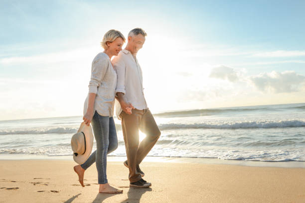 Mature couple walking on the beach at sunset or sunrise. Mature couple walking on the beach at sunset or sunrise. They are having fun, laughing and smiling. The man has grey hair. Ocean in the background. Back lit with lens flare. mature couple stock pictures, royalty-free photos & images