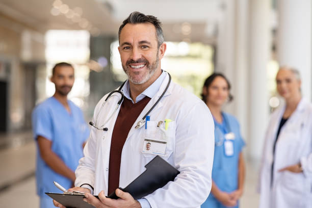 Mature doctor holding medical records at hospital Portrait of handsome mature doctor wearing labcoat and stethoscope reading patient case file in hospital corridor. Successful head physician standing in hospital hallway and looking at camera with his medical team in background. Reliable general practitioner in career with his healthcare staff. Doctor stock pictures, royalty-free photos & images