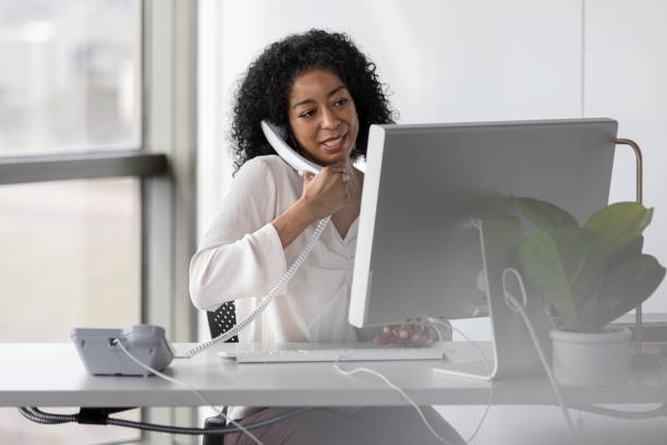 Mature female professor speaks with colleagues while typing on keyboard A mature female professor speaks with colleagues on the landline phone while typing on the keyboard. secretary stock pictures, royalty-free photos & images