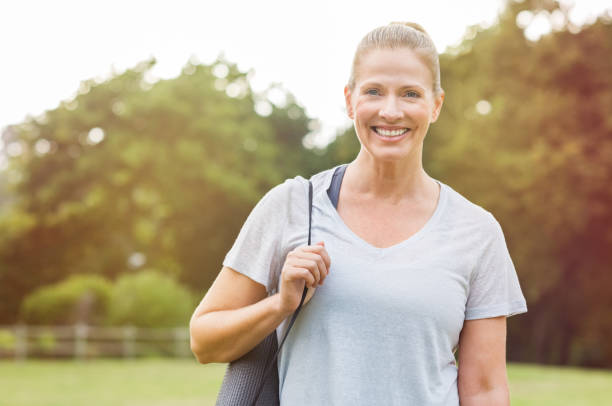 Mature fitness woman Senior beautiful woman holding a grey yoga mat preparing for exercise. Portrait of a mature smiling woman with fitness mat in the park. mid adult women stock pictures, royalty-free photos & images