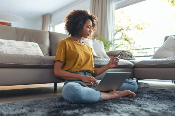 Mature relaxing woman paying something online with her credit card with laptop while sitting on the floor in living room at home. stock photo