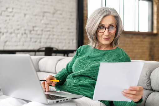 Confident elderly businesswoman is doing paperwork sitting with a laptop on the couch at home, focused senior employee preparing documents, checking bills working remotely at home