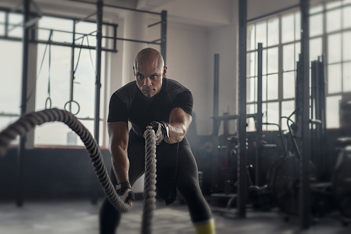 Athlete working out with battle rope at gym. Bald african man training using battle ropes. Fit sportsman doing cross training exercise in an industrial dark gym.