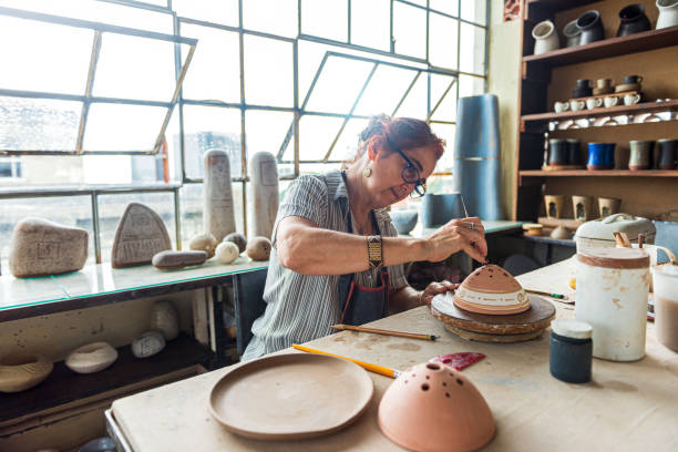 Mature woman artist paints clay bowl Portrait of senior female pottery artist painting a pottery bowl in her workshop art and craft product stock pictures, royalty-free photos & images