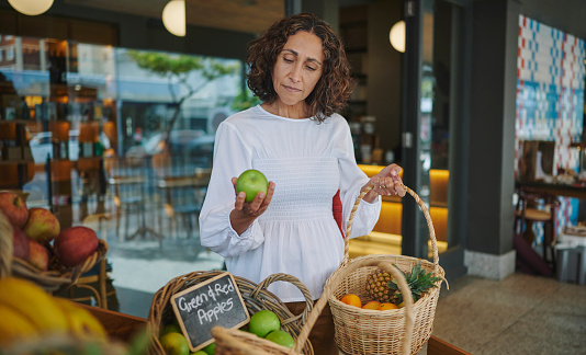 Mature woman with a wicker basket examining an apple while browsing a produce display outside of a supermarket