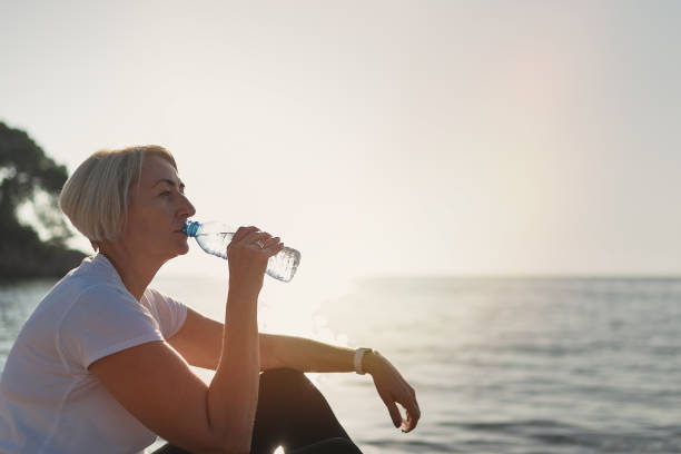 mogen kvinna dricksvatten under solnedgången efter jogging - senior listening music beach bildbanksfoton och bilder