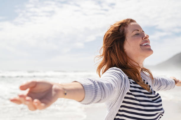 Mature woman enjoy summer beach Happy redhead woman relaxing with arms outstretched at beach on a bright morning with eyes closed. Beautiful middle aged woman felling free at sea. Smiling cheerful mid woman relaxing at the seaside with copy space. carefree stock pictures, royalty-free photos & images