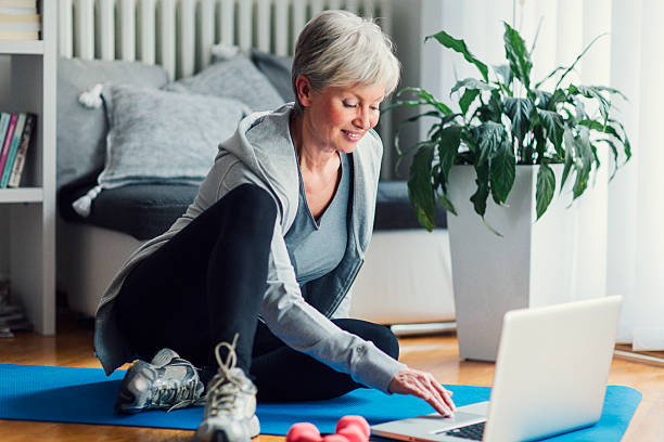 Mature Woman Exercising At Her Home. Mature woman exercising at home, looking for new exercise online on laptop. Sitting on floor and using laptop. Selective focus to her smiling face. russian mature women stock pictures, royalty-free photos & images