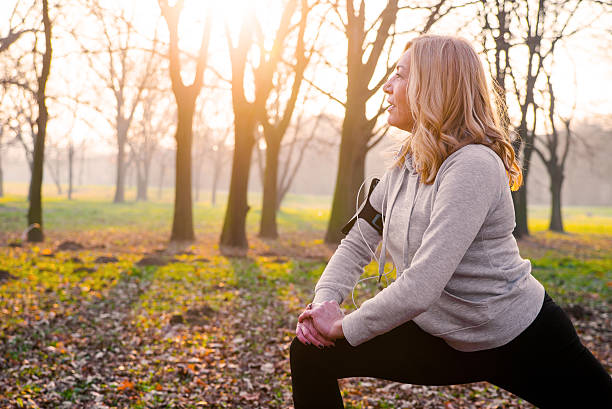 Sportive mature woman l wearing black leggings and gray top doing squatting in autumn nature  holding hands on knee, fitness, copy space.