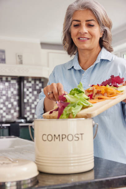 Mature Woman In Kitchen Making Compost Scraping Vegetable Leftovers Into Bin stock photo