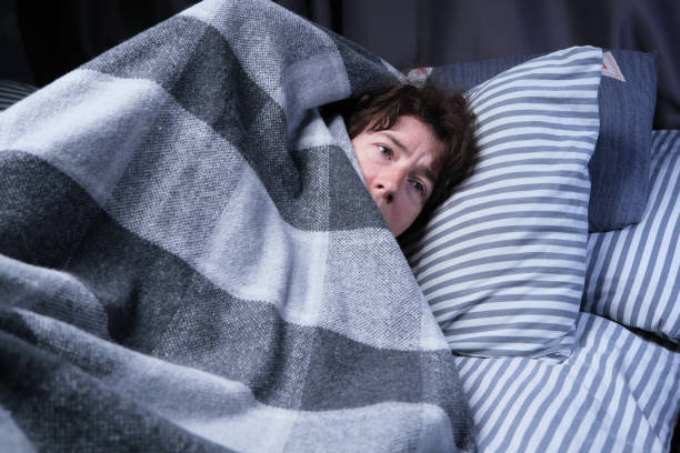 Mature woman is lying in bed but cannot sleep and hiding her head under the blanket stock photo