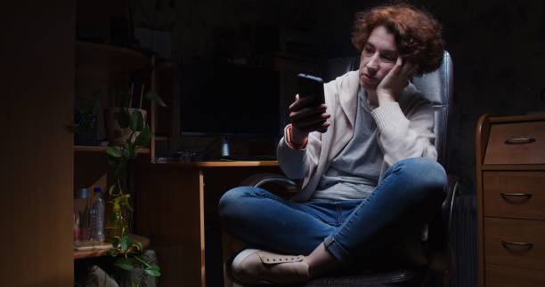 Mature woman is sitting at the desk in dark room and looking at her smartphone in apathy stock photo