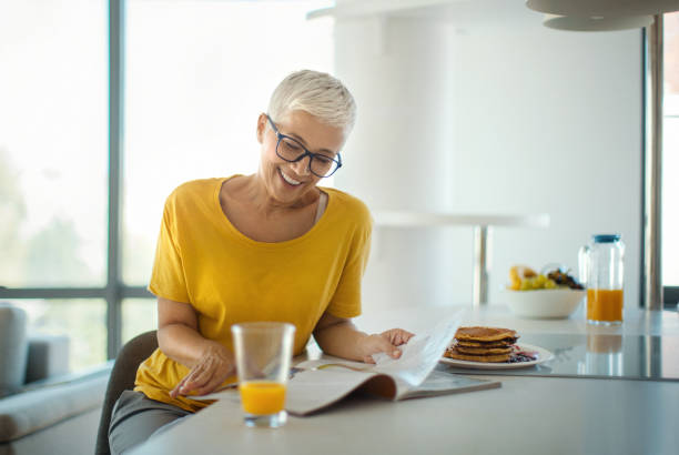 Mature woman reading a magazine while having a breakfast. stock photo