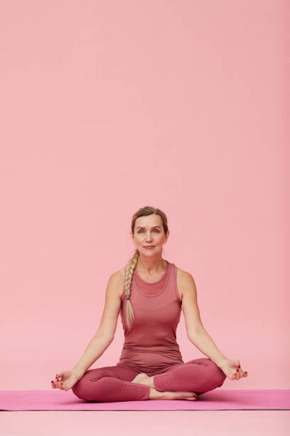 reife frau sitzt in lotus position auf rosa - yoga poses stock-fotos und bilder