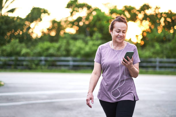 rijpe vrouw die bij zonsondergang loopt die een mobiele telefoon draagt - wandelen lichaamsbeweging stockfoto's en -beelden