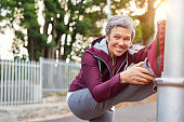 Smiling retired woman listening to music while stretching legs outdoors. Senior woman enjoying daily routine warming up before running at morning. Sporty lady doing leg stretches before workout and looking at camera.