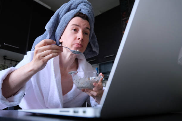 Mature woman wearing bathrobe and hair towel is sitting in domestic kitchen and tasting granola and communicating using laptop stock photo