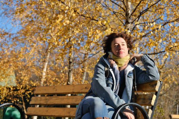 Mature woman wearing warm clothing is sitting on bench on sunny but windy autumn morning stock photo