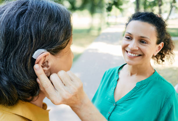 зрелая женщина с нарушениями слуха использует слуховой аппарат для общения со своей подругой на открытом воздухе. слуховые решения - hearing aid стоковые фото и изображения