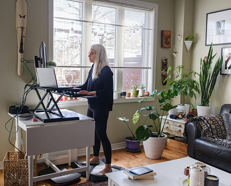 Life in time of COVID-19: Mature woman working on computer from home at the stand up desk. She has long grey hair and dressed in casual black outfit.  Interior of living room set up with home office station, next to window.