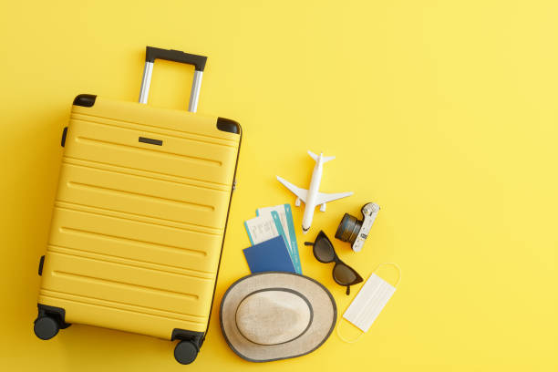 Máscara Médica, Maleta con Sombrero de Sol, Cámara, Pasaporte, Billete de Avión, Gafas de Sol y Avión sobre Fondo Amarillo - foto de stock