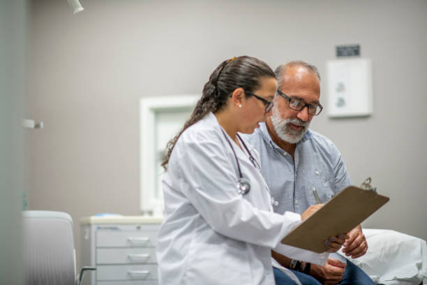 Medical Staff Prepare Their Infectious Control Attire stock photo A female doctor sits next to an elderly male patient during the Covid-19 outbreak.  They are reviewing the patient chart together. medical research stock pictures, royalty-free photos & images