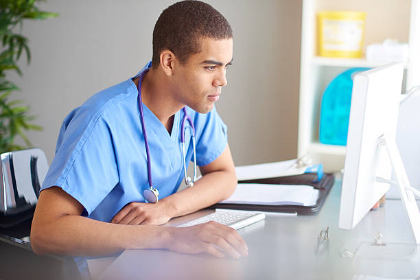 medical student male nurse or doctor sits at his desk looking at his computer screen and writing in his pad . Sunlight pours across the desk from the window beside the desk. He is wearing scrubs . medical student stock pictures, royalty-free photos & images