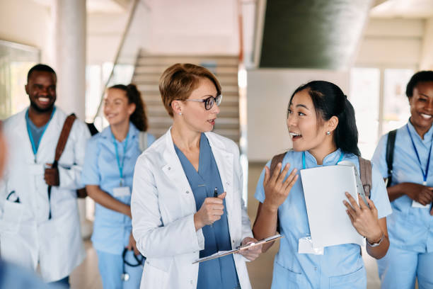 Medical university professor talking to Asian female student in a hallway. Asian nursing student communicating with female doctor while walking with her friends through hallway at medical university. medical schools stock pictures, royalty-free photos & images