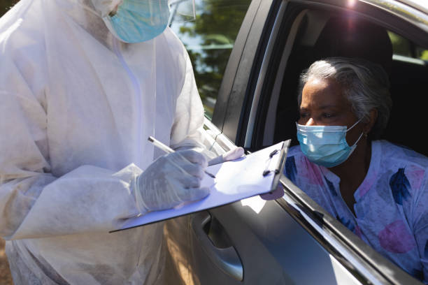 Medical worker wearing ppe suit taking notes talking to senior african american woman sitting in car stock photo