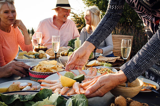 Mediterranean Meal A group of mature friends are sitting around an outdoor dining table, eating and drinking. The shot is a close up of hands setting down a plate of prawns for the meal. tapas stock pictures, royalty-free photos & images