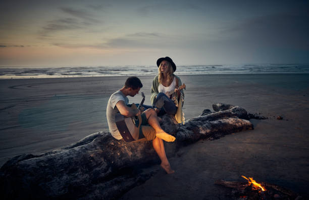 Melodic moments at sunset Shot of a young couple playing guitars together while camping along the coast campfire photos stock pictures, royalty-free photos & images
