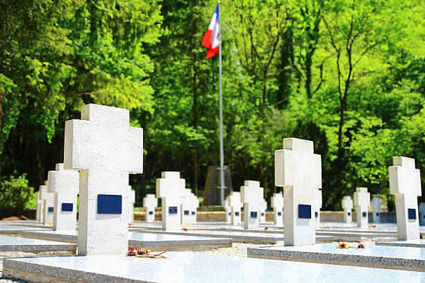 Horizontal composition color photography of white cross shape sculpture shapes in war memorial cemetery with french flag blurred in background due to selective focus on gravestone in foreground. Memorial of maquis of Ain department and of resistance (Memorial des maquis de l'Ain et de la Resistance in french) during second World War (WWII) in small village of Cerdon in Ain, in Bugey mountains in Auvergne-Rhone-Alpes region in France (Europe). This monument of Resistance of Ain and Haut Jura, also named the Valley of Hell (memorial du Val d'Enfer in french), is a memorial attended a memorial and cemetery. This War Memorial, dedicated to the memory of 700 dead maquis of Ain and Haut Jura, will remain a testimony of gratitude to those who gave their lives for our freedom. This cemetery brings 89 graves of resistance fighters or victims of the Shoah; it was inaugurated June 24, 1956 by General de Gaulle. Among them, in addition to local maquis leaders, there are those of foreigners who came to fight in the Ain. Spaniards, Poles, Italians, North Africans lie to their fellow soldiers. Each May 8 ceremony honors all those guerrillas fell for the liberation of France. Picture taken with dried flowers in foreground. Names are not recognizable on gravestones or have been removed, and it's a public place.