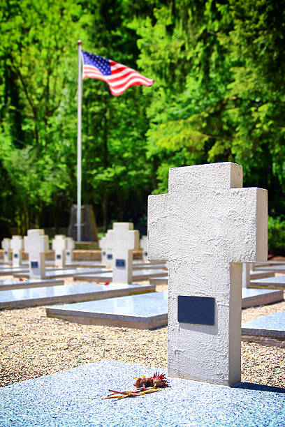 Vertical composition color photography of white cross shape sculpture shapes in war memorial cemetery with USA flag blurred in background due to selective focus on gravestone in foreground. Memorial of Second World War (WWII) in France (Europe). This monument will remain a testimony of gratitude to those who gave their lives for our freedom. Picture taken with a dried flower in foreground and copy space on cross. Names are not recognizable on gravestones or have been removed, and it's a public place.