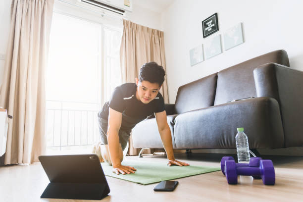 Men workout at home and watch videos for fitness instruction. Asian men in sportswear are exercising in the living room of their homes. stock photo